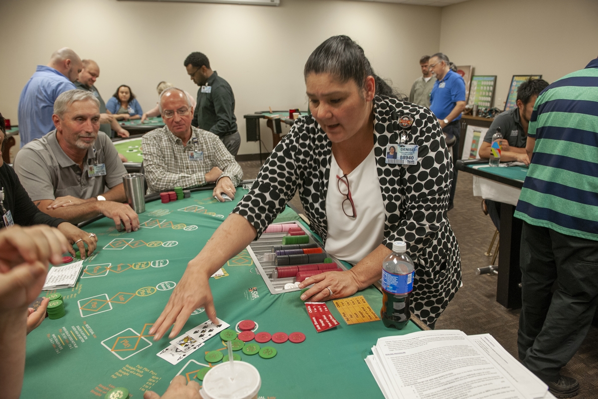 Woman deals a hand of blackjack to several classmates inside a casino training room.