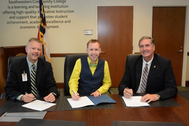 Three men sit at a board room table; man in center signs document.