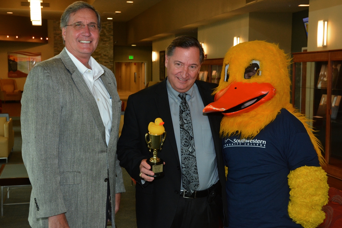 Two men stand beside a life-sized duck mascot. One man holds a duck-topped trophy.