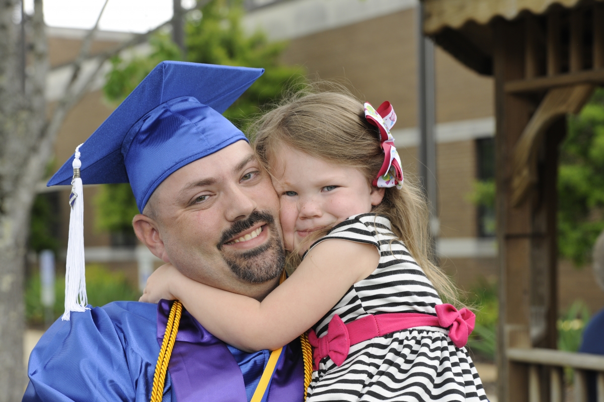 A father, wearing SCC blue cap and gown, holds his daughter outside the college's auditorium.