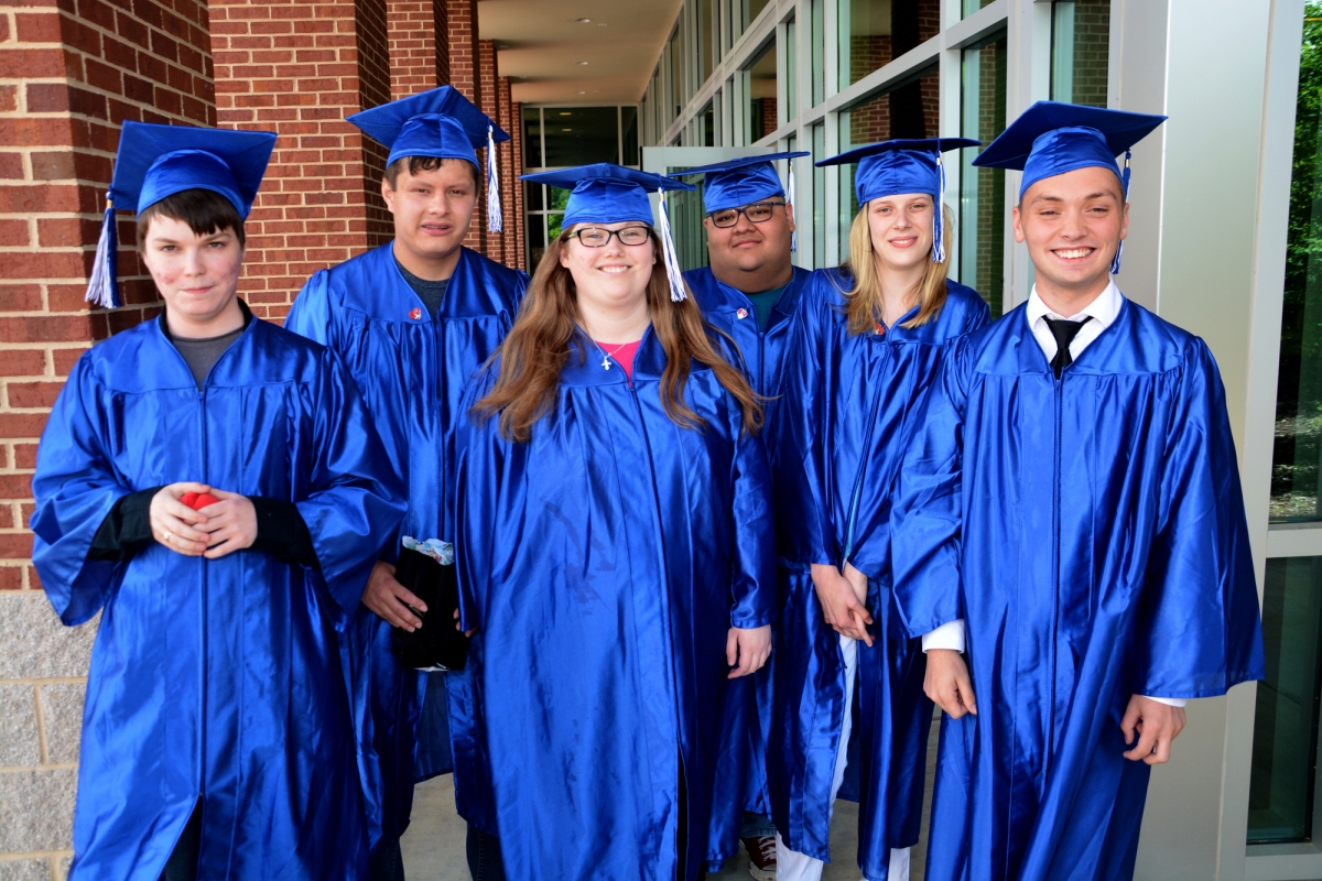 Six graduates wearing blue graduation caps and gowns stand outside a brick buildding
