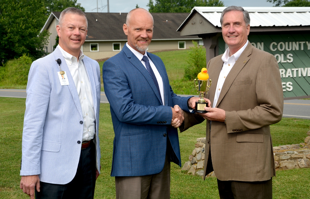 Three men pose outdoors. One handing another a trophy with a rubber duck on it.