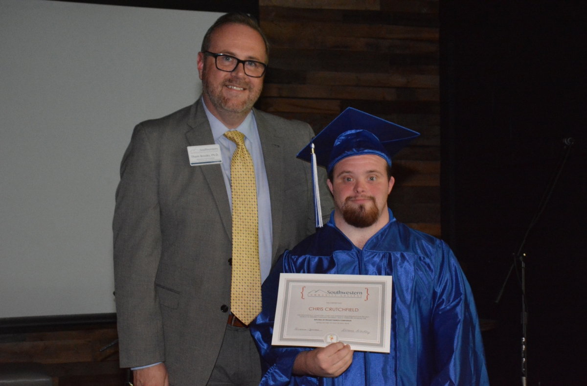 Graduate wearing blue robe and cap stands receives certificate from man wearing suit and tie.