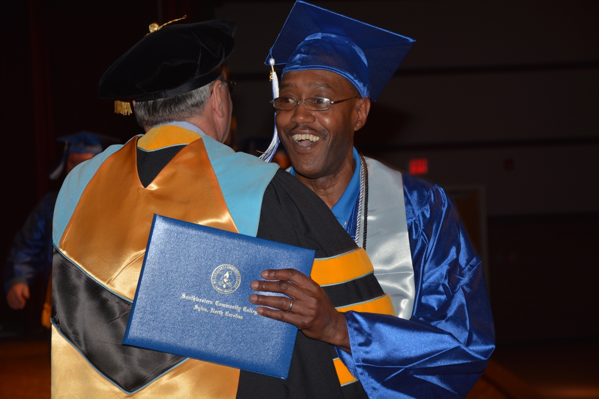 Man wearing blue graduation gown & cap hugs college president after receiving diploma in auditorium.