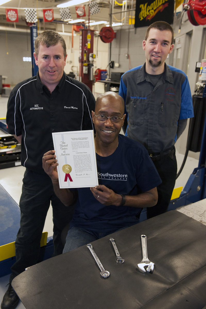 One man holds up a sheet of paper with his US patent; he is flanked by two other men inside SCC's automotiive lab.