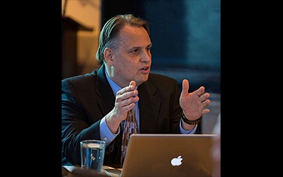 Man, with laptop in front of him, sits at desk and speaks to a group.