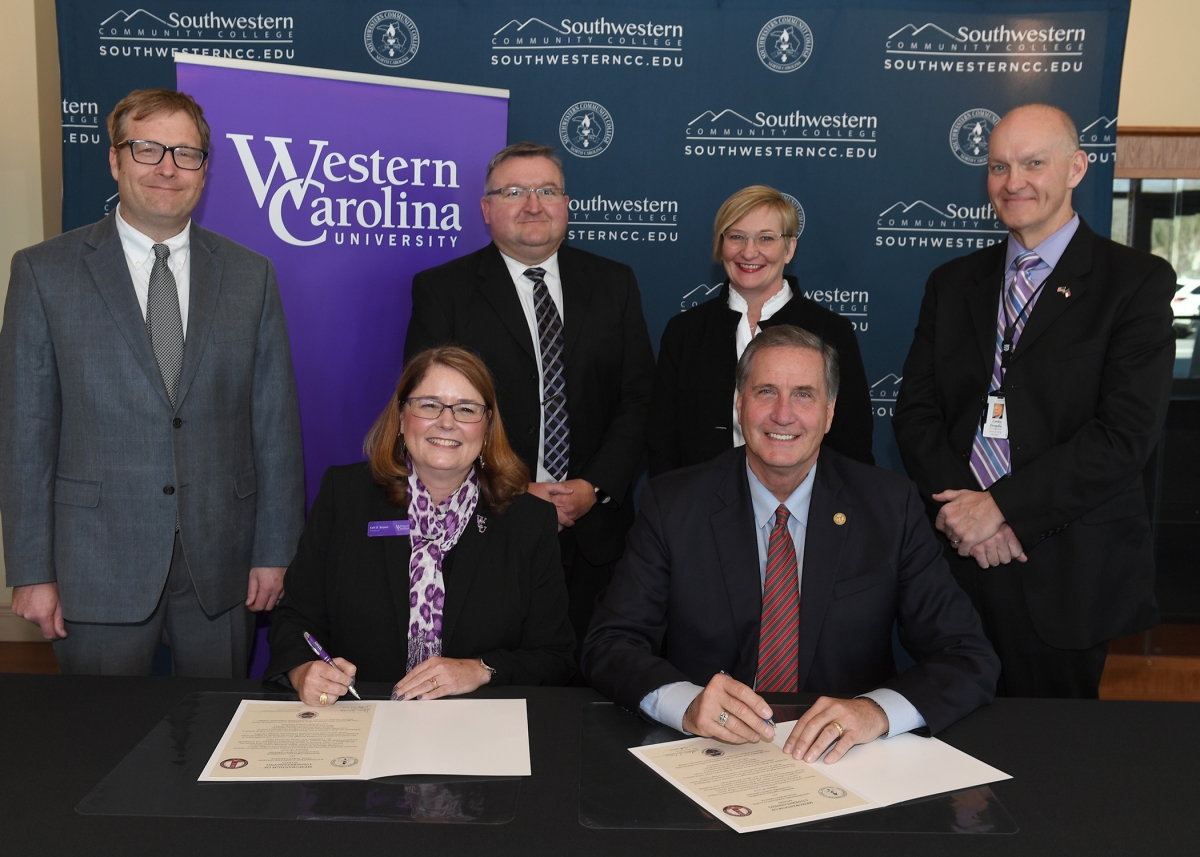 A group of academic leaders gather around in suits and smile as they sign documents