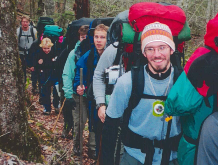 A man smiles and carries a heavy backpack while hiking a trail next to classmates
