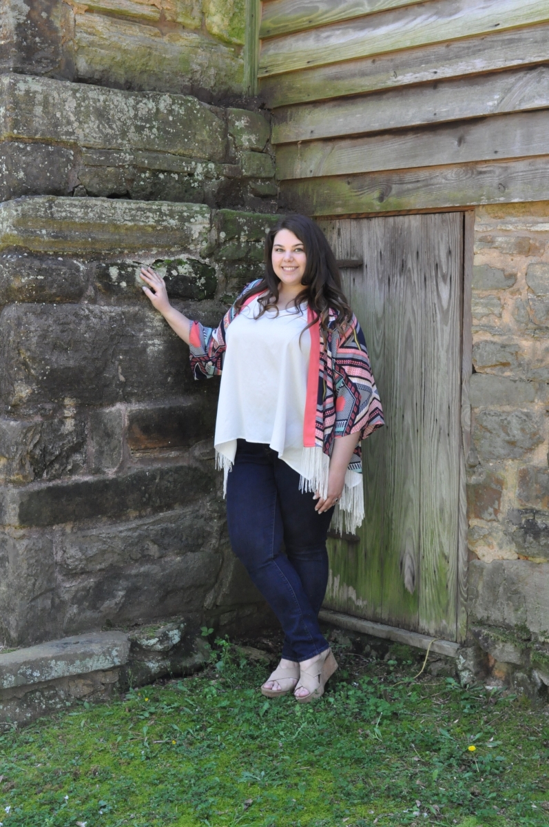 A young woman stands outside of an old wooden building surrounded by nature.