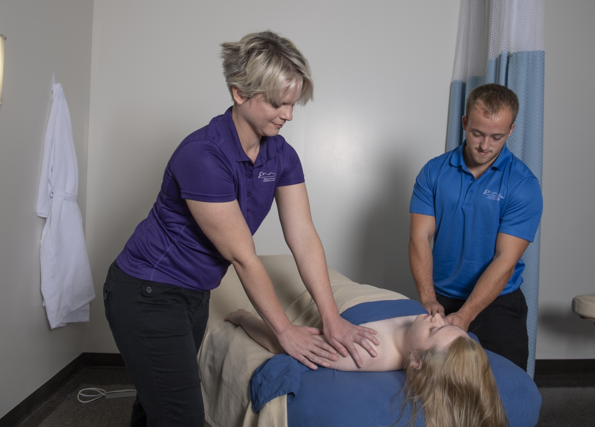 A male and female student gently massage a young woman laying face up on a table.