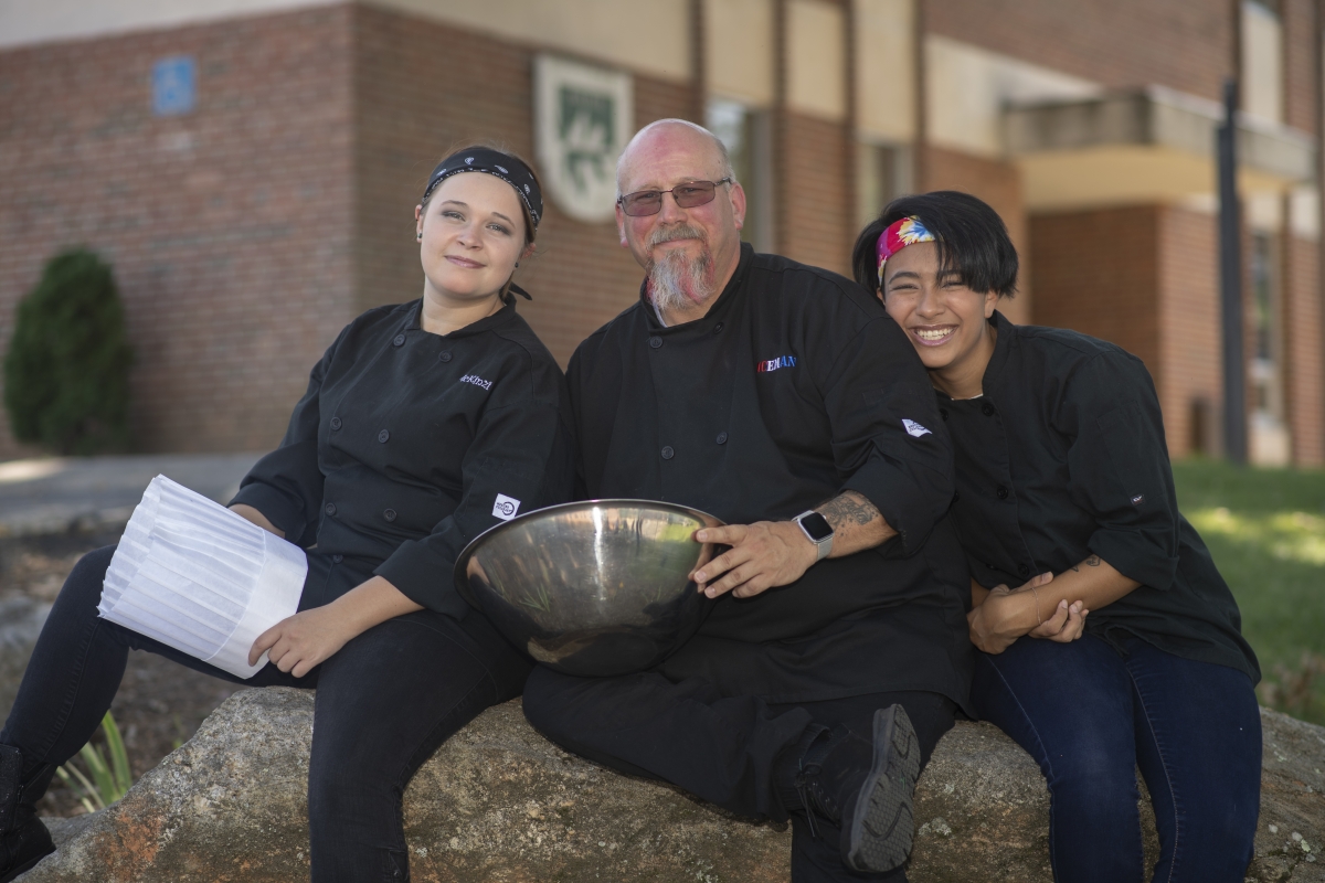 Three culinary students smile in black uniforms while huddled closely outside