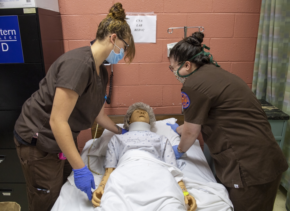 Two females in brown scrubs demonstrate nursing skills on a mannequin