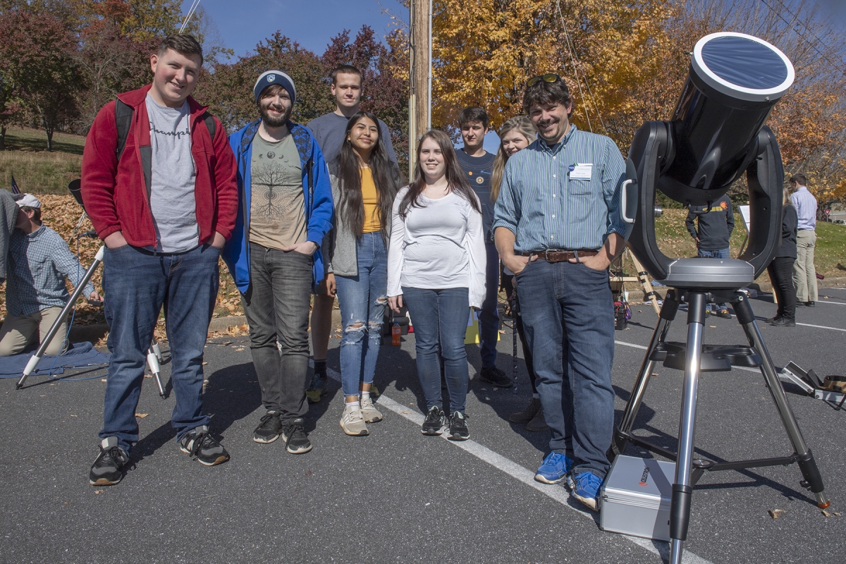 A science instructor and his class smile gathered around a large telescope