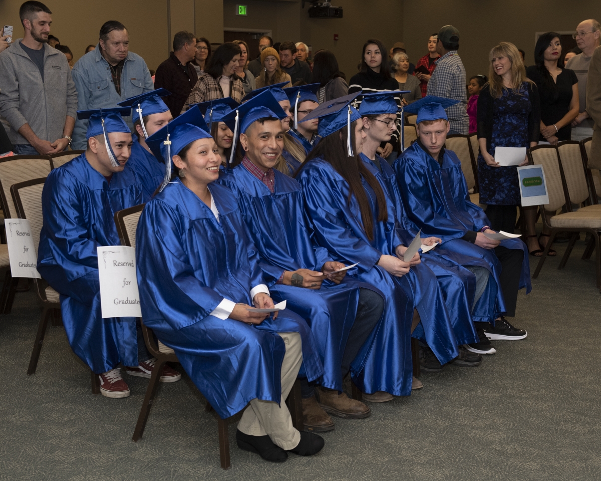 A group of students in blue caps and gowns smile as they sit and wait to receive their diplomas.