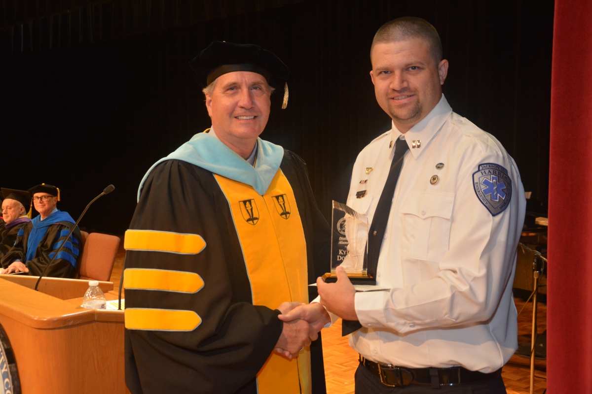 A male college president in a cap and gown presents an award to a male graduate from the emergency medical science program