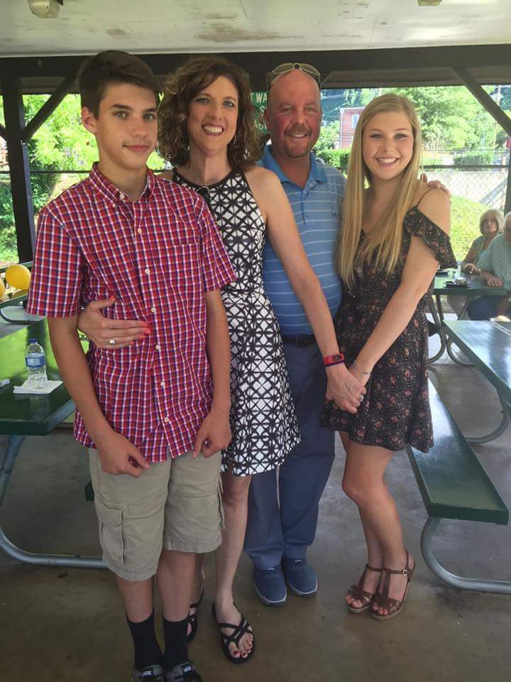 A husband and wife are smiling, posing with their song and daughter on a bright sunny day.