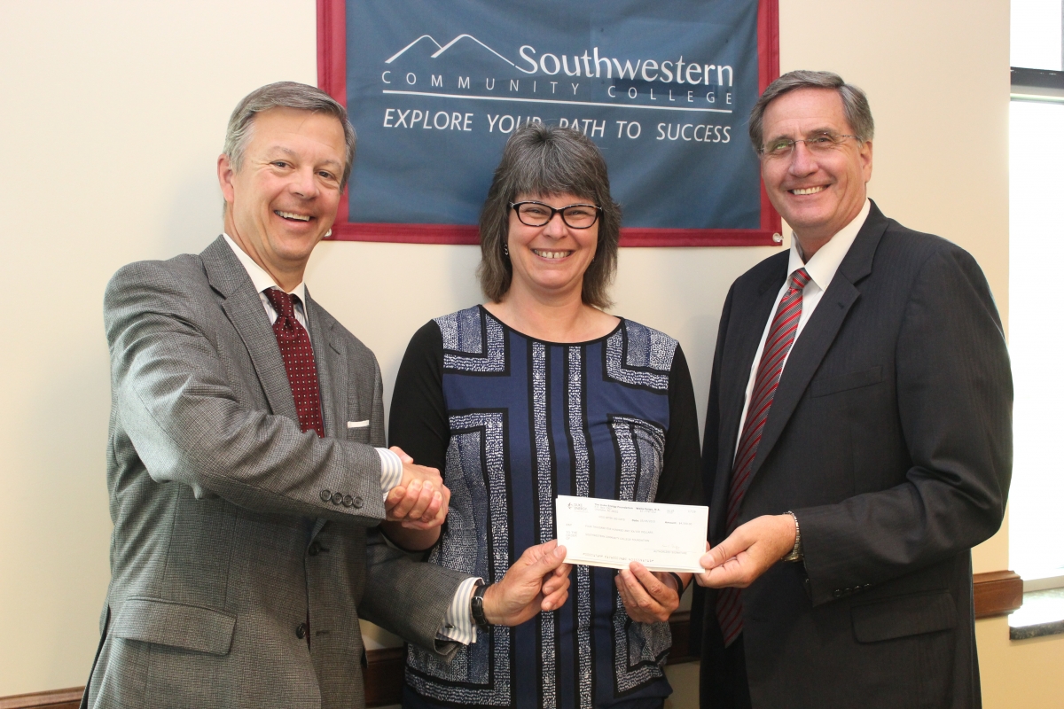 A woman smiles while shaking hands with two men and presenting a check