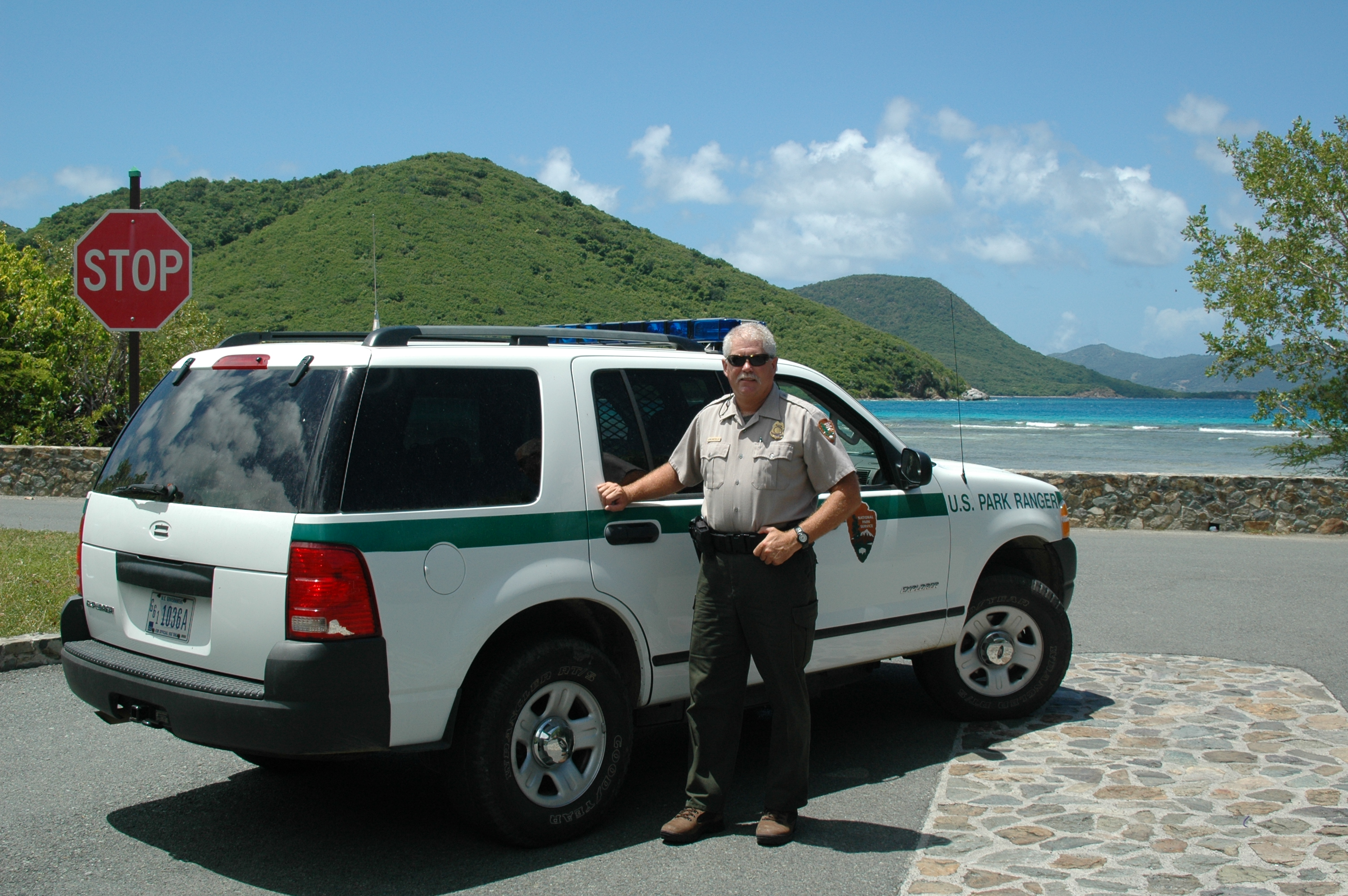 Man wearing NPS uniform stands beside law enforcement vehicle near scenic beach.