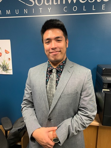 A young man smiles in a gray suit with a floral shirt and silver tie