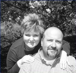 A woman lovingly leans over her husband with her hands placed on his shoulders.