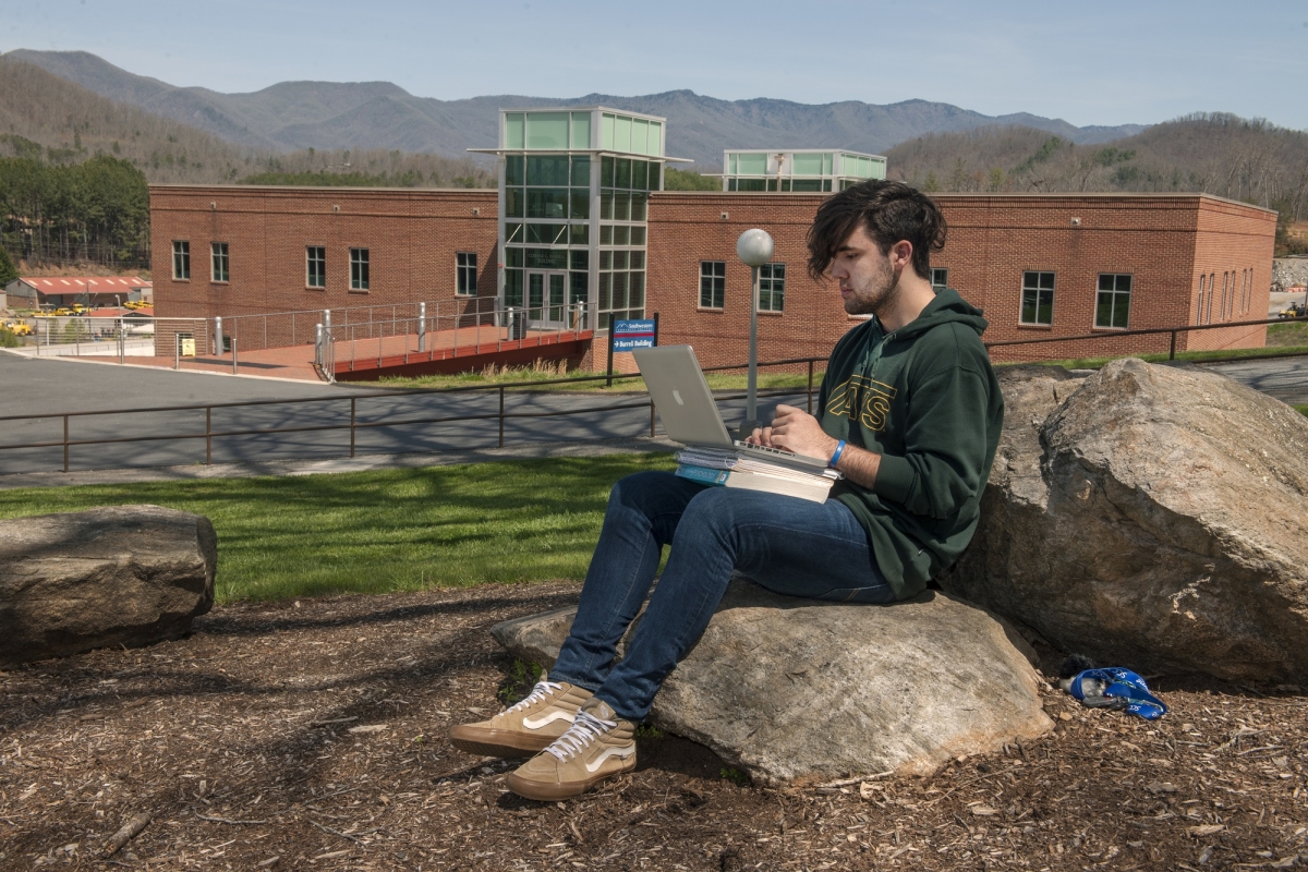 A young man uses a laptop while sitting on a rock outside a school building