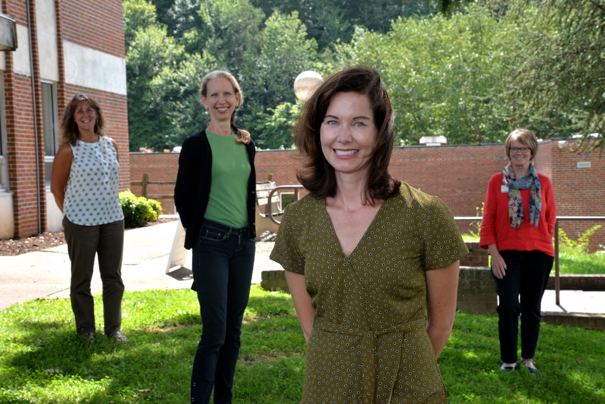 A group of four women stand safely apart while smiling outside