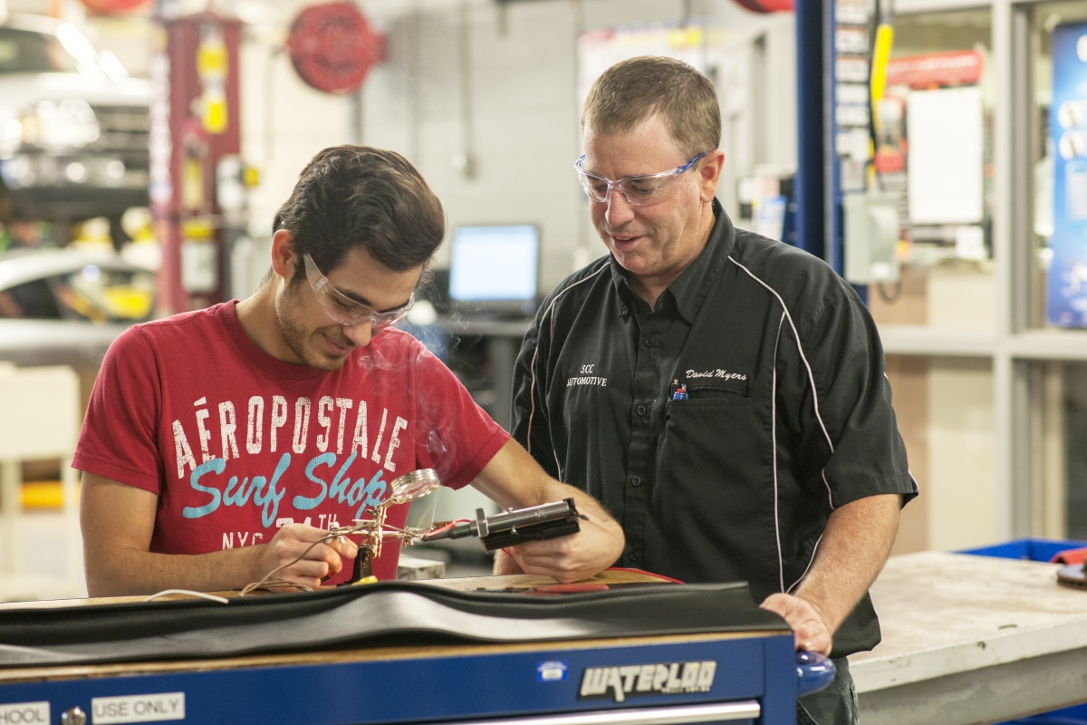 Young man in safety glasses solders two wires together alongside his instructor.