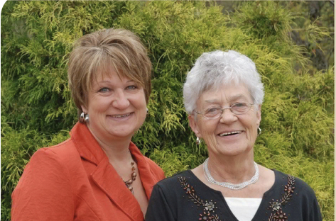 Two women smile and stand side by side outside.