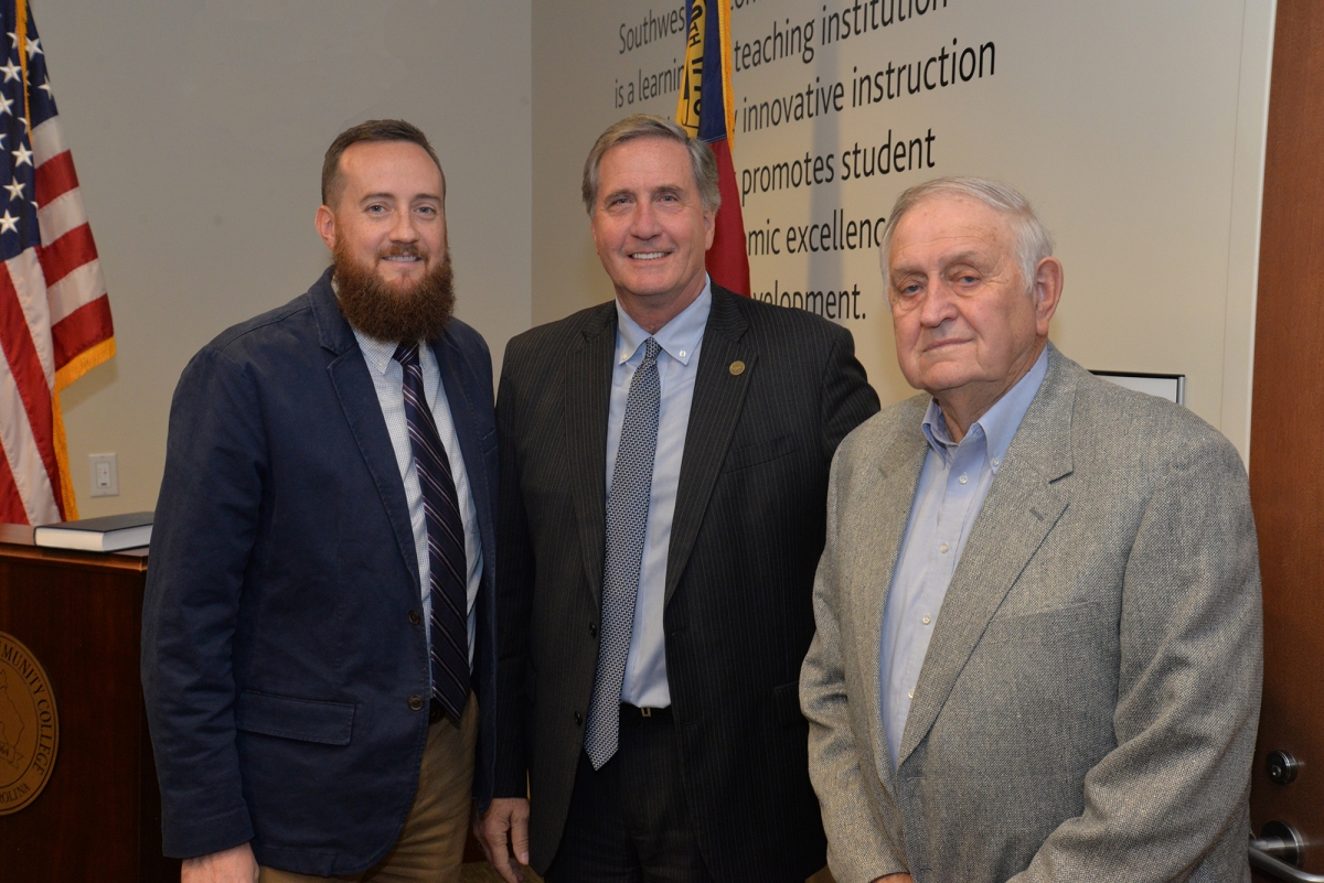 Three men pose in a board room in front of U.S. and North Carolina flags.