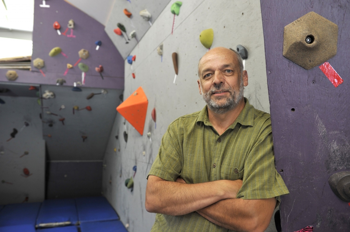 man stands in foreground; indoor rock climbing wall is behind him.