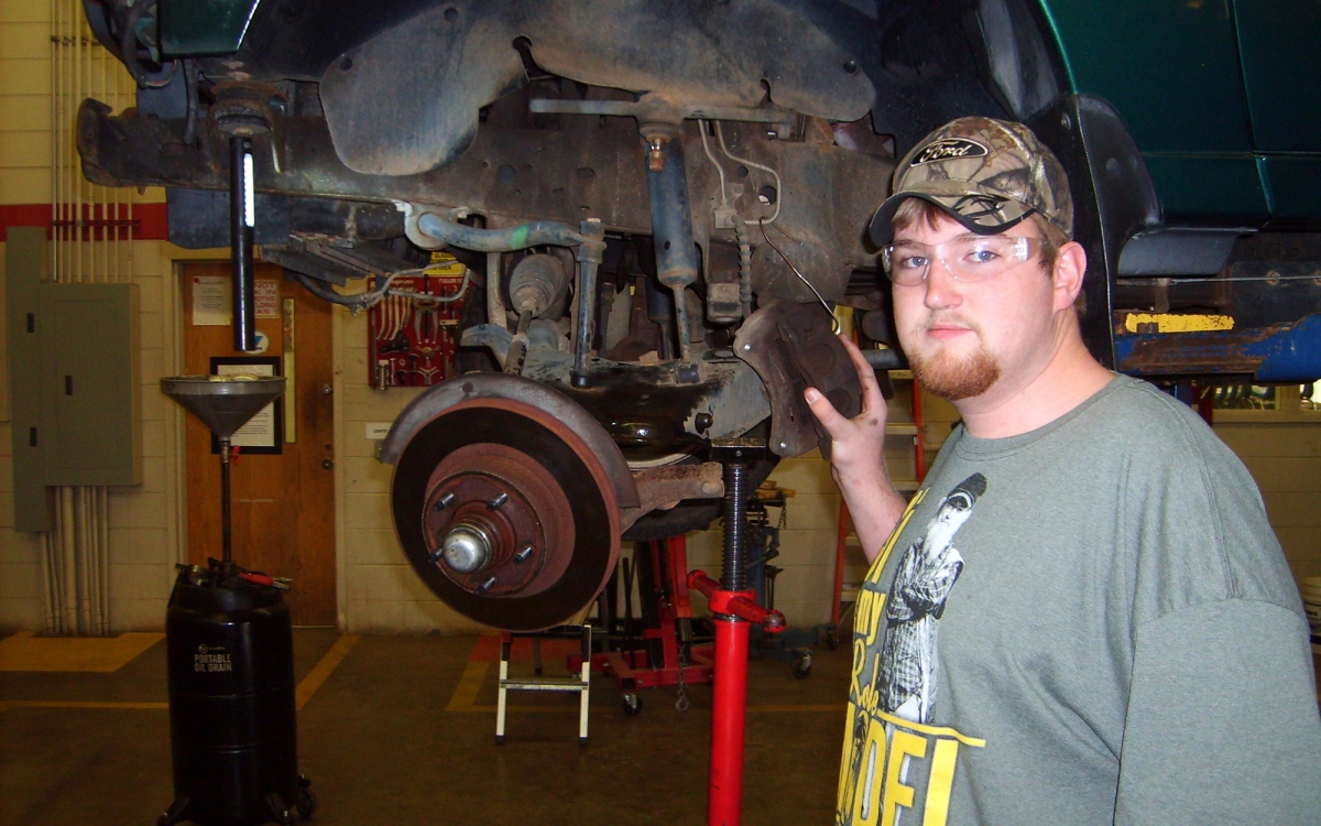 Male student wearing goggles stands beside an automobile that is on a large mechanized jack.