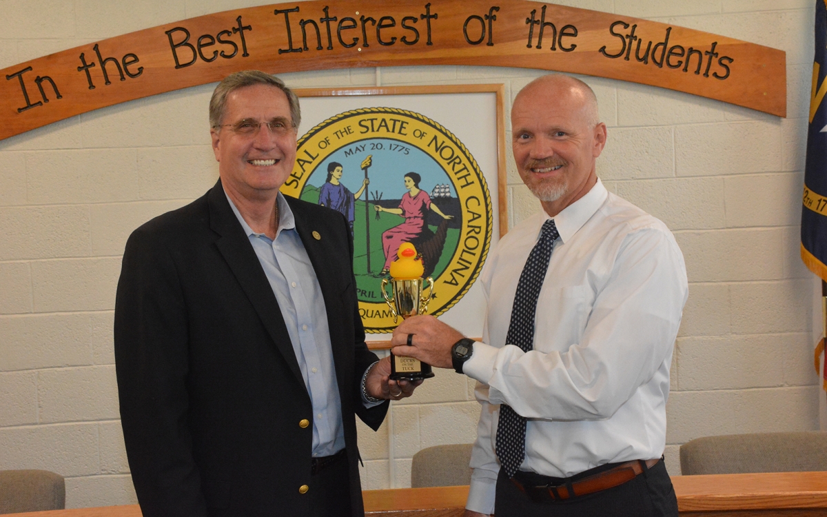 Two men hold a trophy that has a small rubber duck on top. Photo was taken indoors beneath a wall mural that reads 'In the Best Interest of our Students"