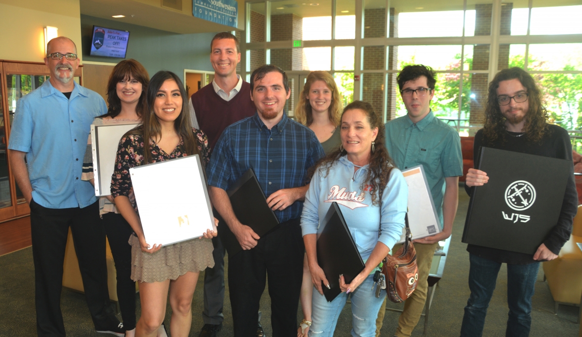 Seven students hold up their portfolios and pose with their instructors