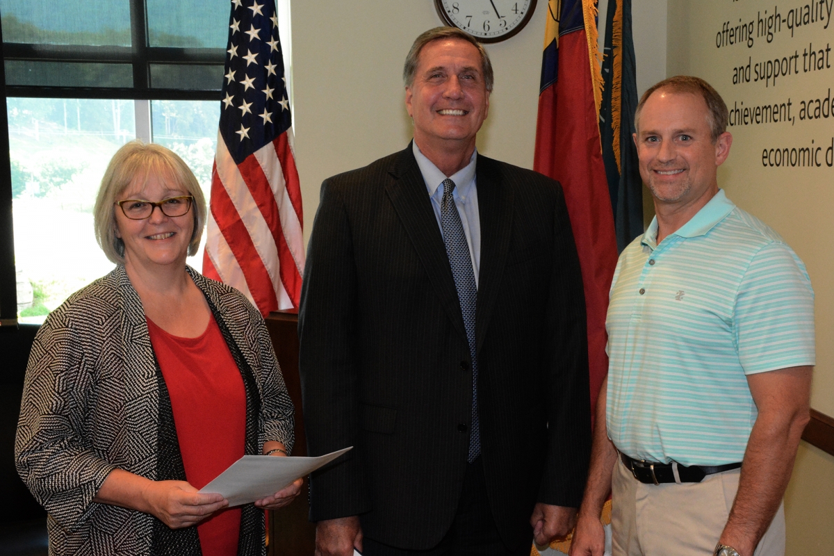 Two men and a woman stand in front of U.S., North Carolina and SCC flags inside a building on SCC's Jackson Campus.