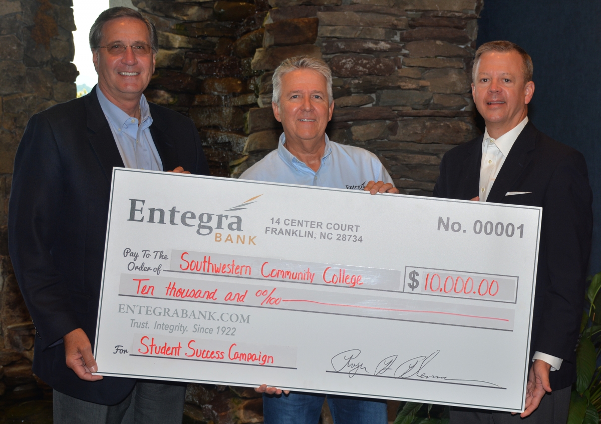 Three men pose with a large check in front of a stacked rock decor