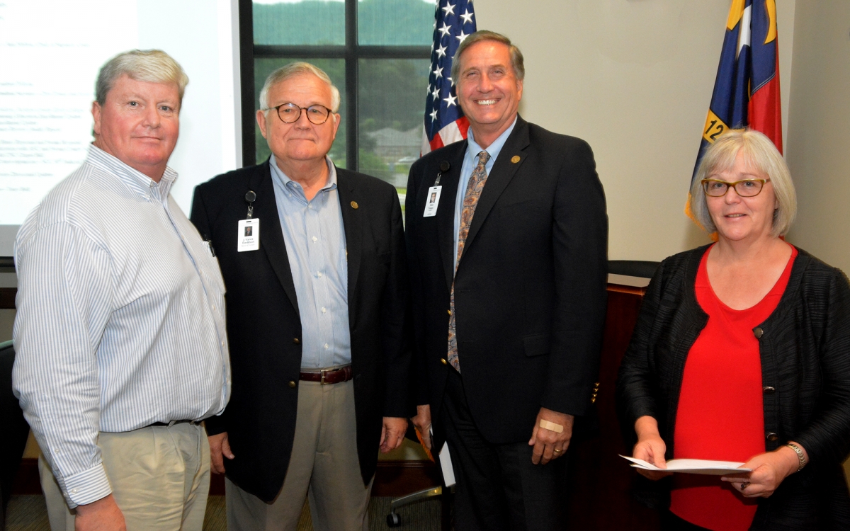 Three men and a woman stand in front of US and NC flags inside a building on SCC's Jackson Campus.