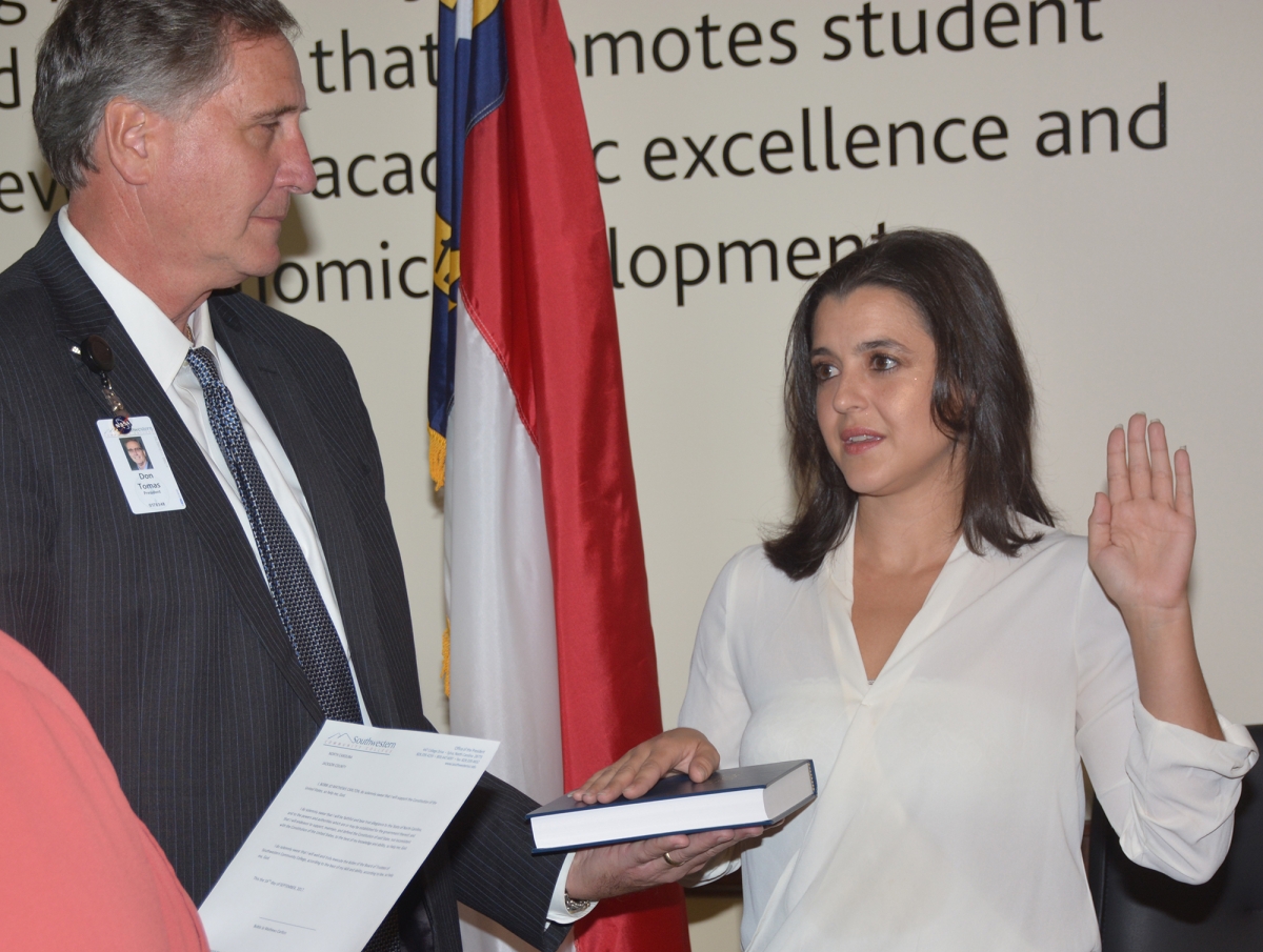 Woman places one hand on a bible and holds the other arm in the air as she's sworn in. Man holds the bible for her.