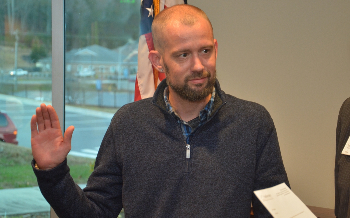 Standing in front of a U.S. flag, a man holds up one hand and places the other on the Bible.