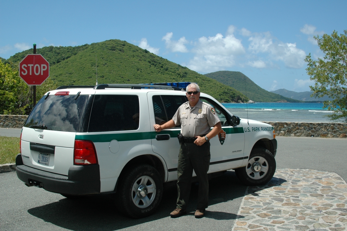 Man wearing NPS uniform stands beside a law enforcement vehicle near a scenic beach