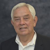 Man in white shirt and suit coat stands in front of gray background.