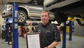 Man holds plaque in garage with cars on lifts in background.