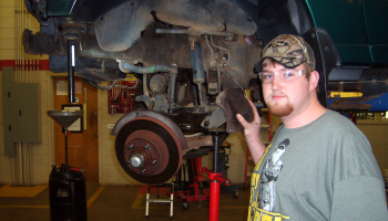 Male student wearing goggles stands beside an automobile that is on a large mechanized jack.