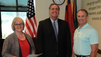 Two men and a woman stand in front of U.S., North Carolina and SCC flags inside a building on SCC's Jackson Campus.