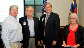 Three men and a woman stand in front of US and NC flags inside a building on SCC's Jackson Campus.