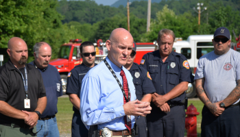 Curtis Dowdle speaks; first responders and emergency vehicles are in the background.