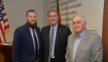 Three men pose in a board room in front of U.S. and N.C. flags.