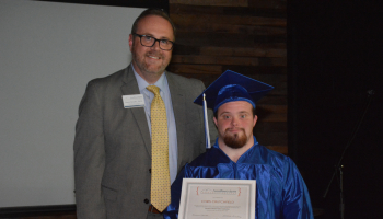 Graduate wearing blue robe and cap stands receives certificate from man wearing suit and tie.