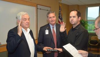 Two men hold up their right hands and place left hands on bible held by a third man. A fourth man reads pledge from a sheet of paper.