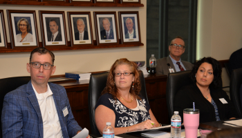 Three employees sitting at board room table listen to a presentation.