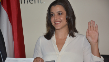 Woman places one hand on a bible and holds the other arm in the air as she's sworn in. Man holds the bible for her.